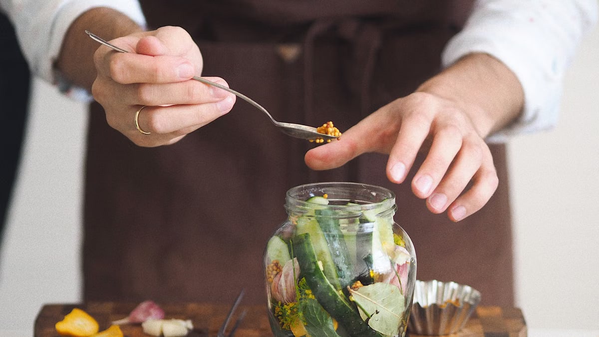 Close-up of hands preparing homemade pickles with fresh vegetables in a glass jar on a wooden board.